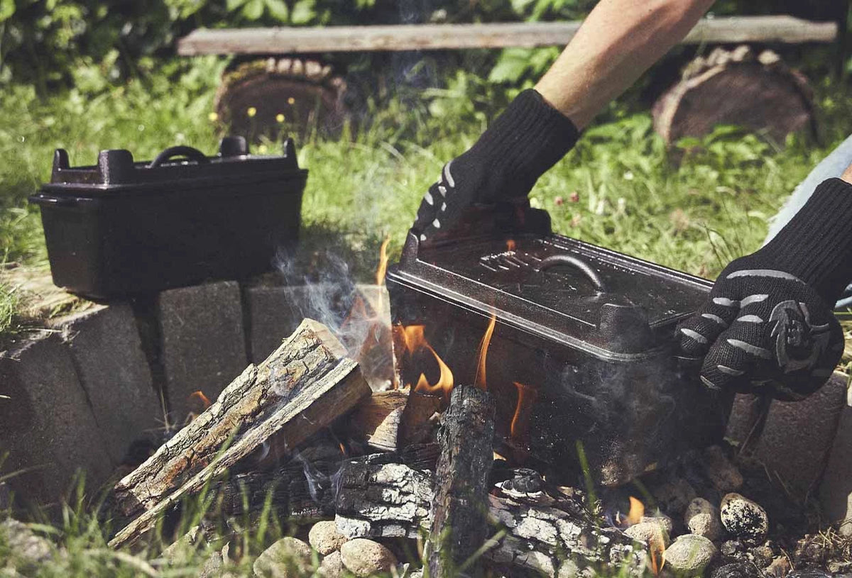 Lagerfeuerszene Kastenform wird mit Handschuhen an den Griffen von den Holzscheiten gehoben Lagerfeuerszene Kastenform wird mit Handschuhen an den Griffen von den Holzscheiten gehoben