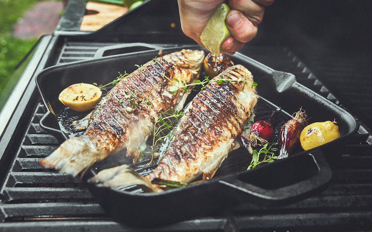 Grillszene Fisch wird in Grillpfanne auf Grill gegart und mit Zitronensaft beträufelt Grillszene Fisch wird in Grillpfanne auf Grill gegart und mit Zitronensaft beträufelt
