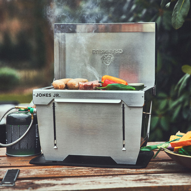 Burnhard JONES Jr. 1-Brenner Klappgrill mit Würstchen und Grillgemüse auf einem Picknick Tisch Burnhard JONES Jr. 1-Brenner Klappgrill mit Würstchen und Grillgemüse auf einem Picknick Tisch