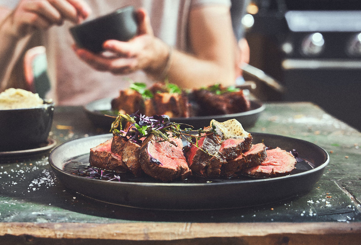 Image 35870143643963 Tischszene Teller mit ästhetisch angerichtetem Fleisch im Hintergrund werden Gewürze aus Dipschale entnommen