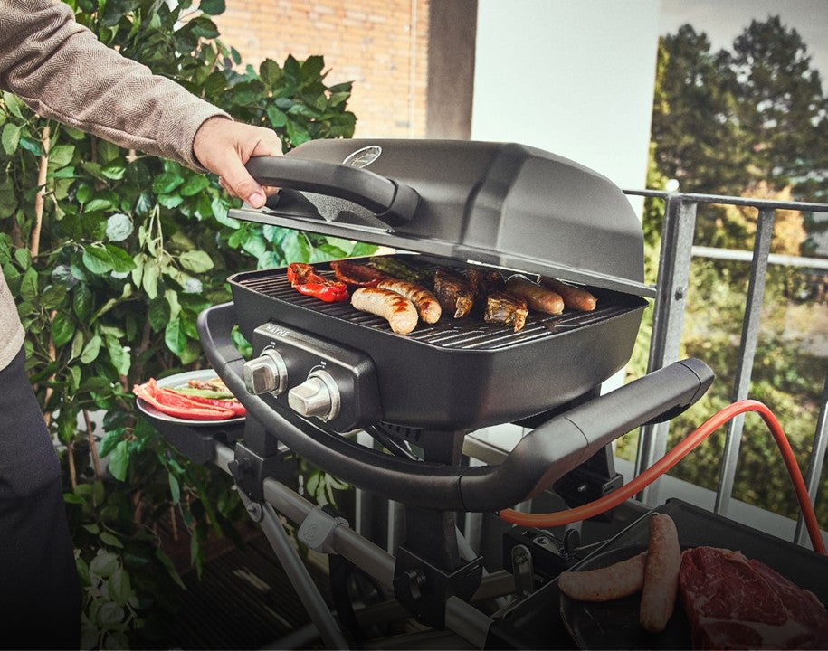 WAYNE Gasgrill auf Balkon wird mit der Hand geöffnet, Würstchen & Grillgut auf Grillrost