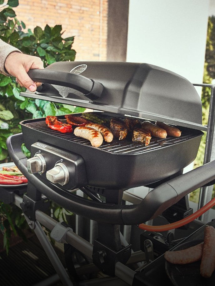 WAYNE Gasgrill auf Balkon wird mit der Hand geöffnet, Würstchen & Grillgut auf Grillrost