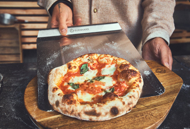 Fertig gebackene Pizza wird mithilfe der Schaufel auf dem Pizzabrett abgelegt