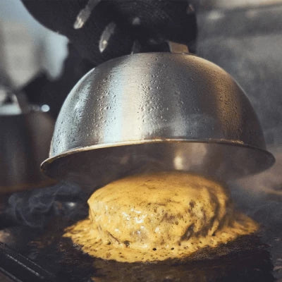 Grillszene Burgerglocke wird auf Grillplatte angehoben darunter Patty mit schmelzender Käsescheibe