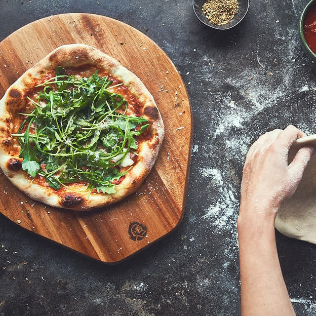 Draufsicht Tischszene Pizzabrett mit fertiger Pizza und daneben wird Pizzateig ausgerollt